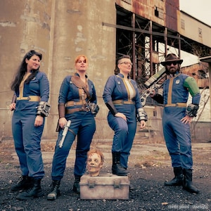 May include: Four individuals wearing blue jumpsuits with yellow trim, reminiscent of Vault-Tec uniforms. They are equipped with various props including wrist-mounted devices, a silver pistol, a rifle, and a severed head prop on a metal box.