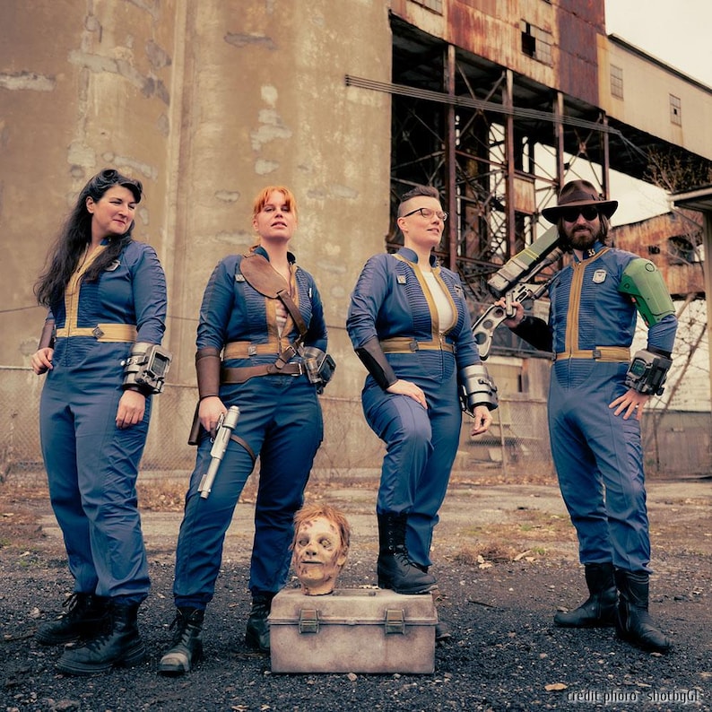 May include: Four individuals wearing blue jumpsuits with yellow trim, reminiscent of Vault-Tec uniforms. They are equipped with various props including wrist-mounted devices, a silver pistol, a rifle, and a severed head prop on a metal box.
