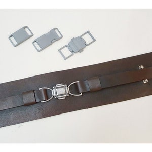 May include: A brown leather belt with a silver buckle. The belt has two straps and is secured with a silver buckle. Three gray plastic buckles are also included.