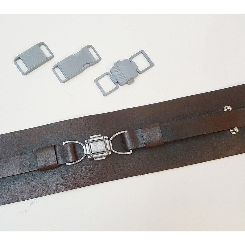 May include: A brown leather belt with a silver buckle. The belt has two straps and is secured with a silver buckle. Three gray plastic buckles are also included.
