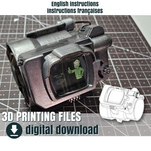 Op de afbeelding: Een metallic grijze prop met een scherm waarop een groene Vault-Tec-mascotte te zien is die een duim omhoog geeft. De tekst "English instructions instructions françaises" en "3D PRINTING FILES digital download" is zichtbaar.