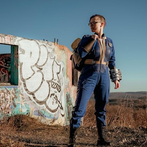 May include: A blue jumpsuit with a tan vertical zipper panel and collar, worn with a tan backpack, black boots, and a metallic forearm gauntlet. The outfit features a dark blue quilted texture on the chest and shoulders, paired with black glasses.