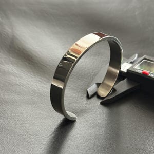 Könnte beinhalten: Ein poliertes silbernes Armband mit flachem, rechteckigem Design. Das Armband wird auf einer schwarzen Oberfläche neben einem digitalen Messschieber gezeigt. Das Armband ist etwa 1,3 cm breit.