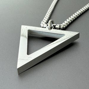May include: A silver pendant necklace with a geometric triangle design. The pendant is a hollow, three-dimensional triangle suspended from a box chain. The chain and pendant are silver. The background is a solid grey.