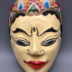 May include: A hand-painted, cream-colored wooden mask with a serene expression. The mask features black eyebrows and eyes, a red mouth, and a decorative headdress with gold, red, blue, and yellow accents. The mask is likely for theatrical or decorative purposes.