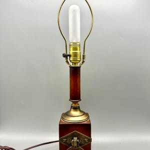May include: A vintage table lamp with a dark wood base and a brass-colored metal accent. The lamp features a long, cylindrical light bulb and a decorative handle on the front. The lamp's cord is brown.