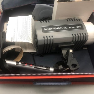 May include: A gray and black Model PG4001-ML AC 110-120V studio light is open inside a red and blue zippered case. The case contains a light stand, paperwork, and other accessories. The light stand is labeled AE41 FUS.