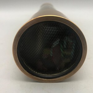 May include: A bronze kaleidoscope with a black mesh lens. The kaleidoscope has a circular lens and a cylindrical body. The lens is surrounded by a bronze ring. The body of the kaleidoscope is bronze in colour.