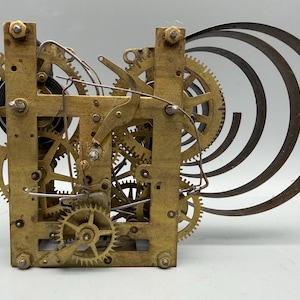 May include: A brass clock movement mechanism featuring an intricate arrangement of interlocking gears, wheels, and metal plates. Attached to the side are several curved, dark metal chime rods extending outward from the central frame structure.