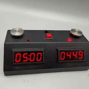May include: A black digital chess clock with two red LED displays showing 05:00 and 04:49. The device features two round silver buttons on the top, a small red power button in the center, and two small red indicator lights above the displays.