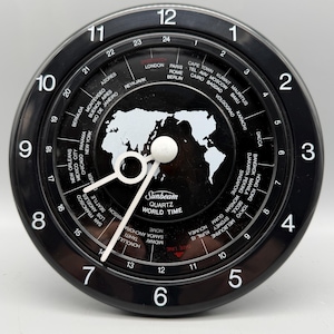 May include: A black Sunbeam quartz world time clock with a white map graphic on the face. The dial features large white numbers 1 through 12, a 24-hour inner ring, and a list of global cities. White hour, minute, and second hands indicate the time.