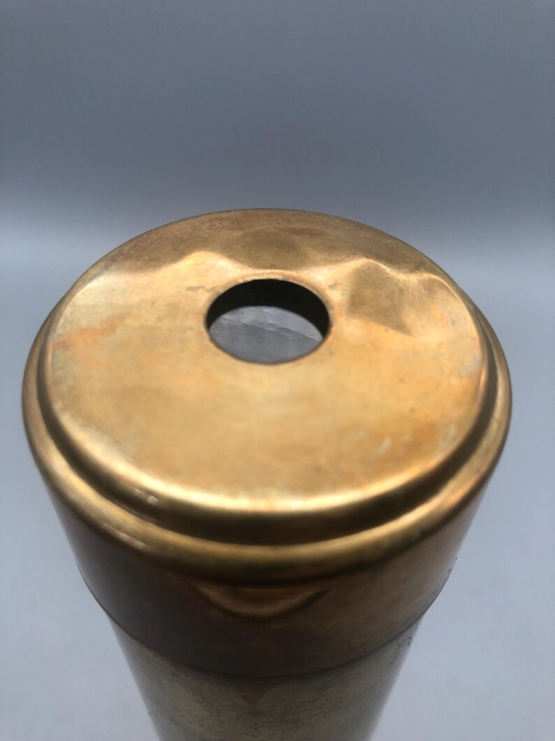 May include: Close-up of a brass kaleidoscope with a circular viewing hole. The brass has a warm, golden hue and shows signs of age. The body of the kaleidoscope is cylindrical, with a darker band near the base.