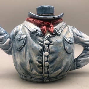 May include: A novelty teapot shaped like a denim jacket, cowboy hat, and red neckerchief. The teapot is painted in shades of blue and white, with button and pocket details. A unique and decorative kitchen item.