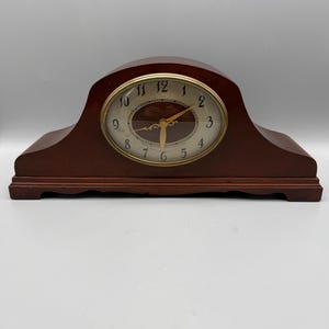 May include: A dark brown wooden mantel clock with a curved top and a rectangular base. The clock face is circular with a gold-colored rim, featuring black Arabic numerals, gold-colored hands, and a dark brown oval center detail.