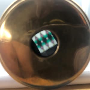 May include: A shiny, gold-coloured metal ashtray with a central, circular inset. The inset features a black background with a diamond pattern in green, white, and pink. The ashtray has a smooth, reflective surface.