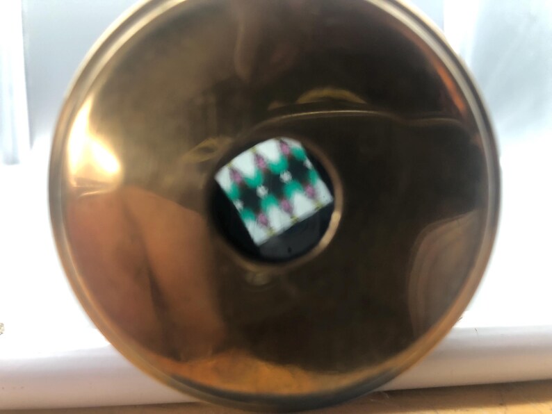 May include: A shiny, gold-coloured metal ashtray with a central, circular inset. The inset features a black background with a diamond pattern in green, white, and pink. The ashtray has a smooth, reflective surface.