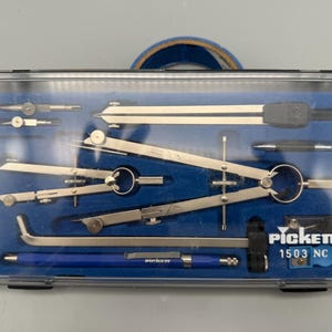 May include: A Pickett 1503 NC drafting set in a clear plastic case with a blue foam insert. The set includes various metal compasses, a divider, a metal extension bar, and a blue mechanical pencil, all organized in custom-fitted slots.