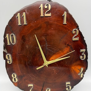May include: A wall clock featuring a circular, polished dark brown wood slice as the face. The clock has gold-colored metal numerals from 1 to 12 arranged around the perimeter and three thin, gold-colored metal hands at the center.