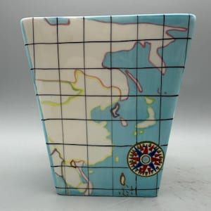 May include: A ceramic vase with a world map design. The vase is a light blue color with a white background. The map features black grid lines and a compass rose. The vase has a rectangular shape.