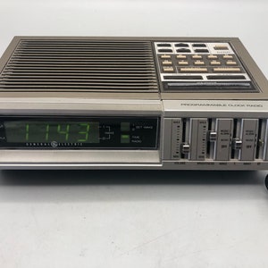 May include: A vintage General Electric programmable clock radio. The digital display shows 11:43 AM. The device has a silver and gray color scheme with buttons for radio and alarm settings. It features volume, brightness, and wake controls.