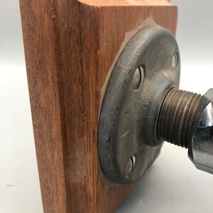 May include: A wooden base with a metal pipe fitting attached to it. The base is square and has a dark brown finish. The pipe fitting is grey and has a threaded end.