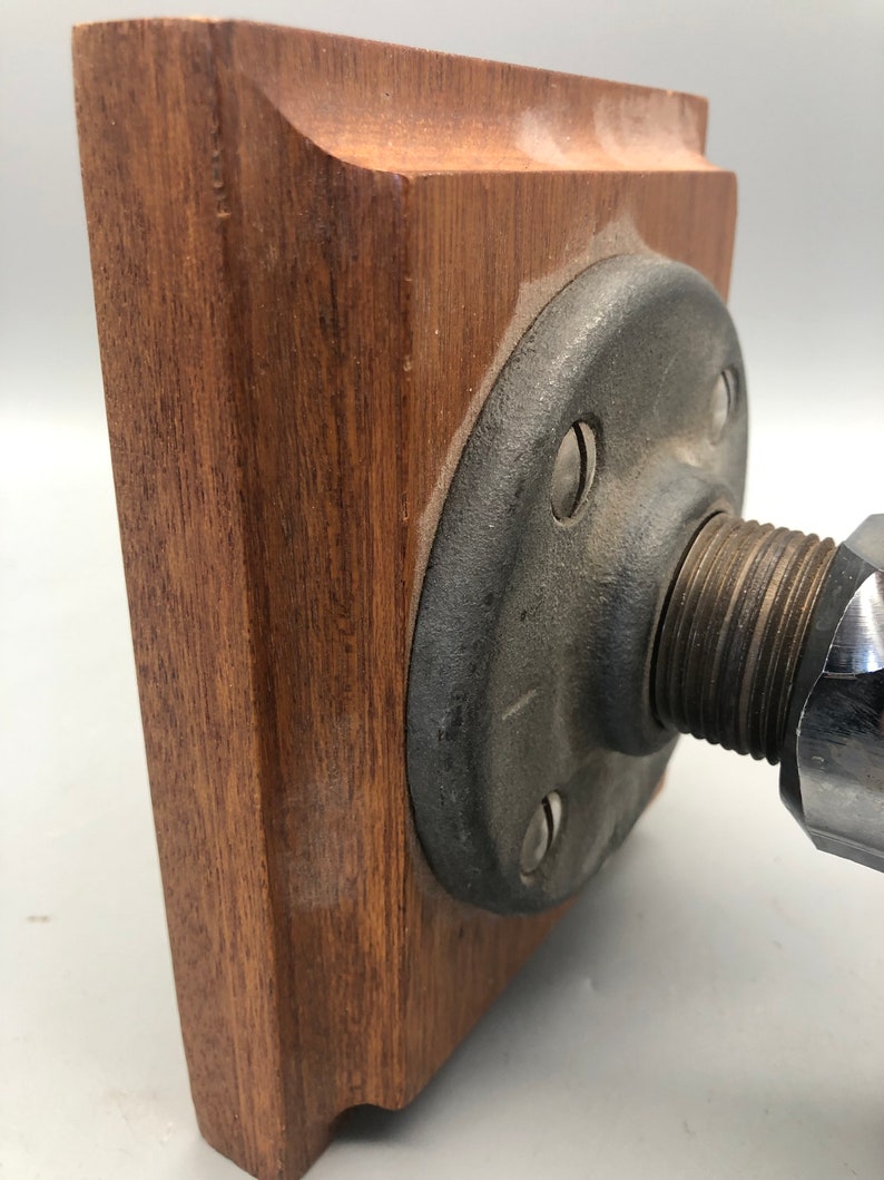 May include: A wooden base with a metal pipe fitting attached to it. The base is square and has a dark brown finish. The pipe fitting is grey and has a threaded end.