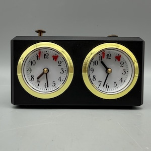 May include: A black analog chess clock with two circular white clock faces, each featuring black numerals and gold-toned bezels. Two small red indicators are positioned near the top of each dial, and two metal buttons are located on the top.