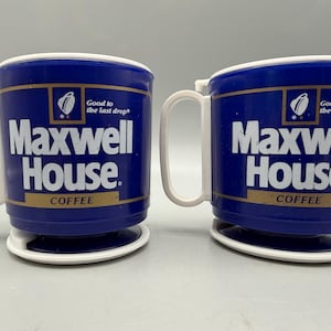 Könnte beinhalten: Zwei blaue Kunststoffbecher mit weißen Henkeln und Sockeln. Jeder Becher zeigt den weißen Schriftzug Maxwell House, den Slogan Good to the last drop und eine Kaffeetassengrafik. Die Becher haben ein goldenes Querband und einen weißen oberen Rand.