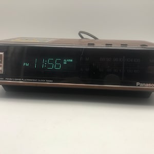 May include: A brown Panasonic FM-AM 2-band fluorescent clock radio with a digital display showing 11:56 PM. The radio has a step dimmer and an alarm function.