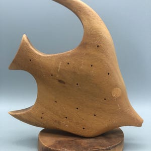 May include: A wooden sculpture of a fish with a curved dorsal fin, mounted on a round wooden base. The fish is light brown with small drilled holes and a circular marking. The sculpture has a mid-century modern aesthetic.