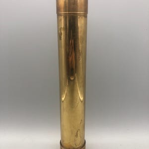 May include: A cylindrical brass container with a removable lid and base. The container has a polished, reflective surface with vertical lines. The lid and base are a darker shade of brass. The container is approximately 25 cm tall.