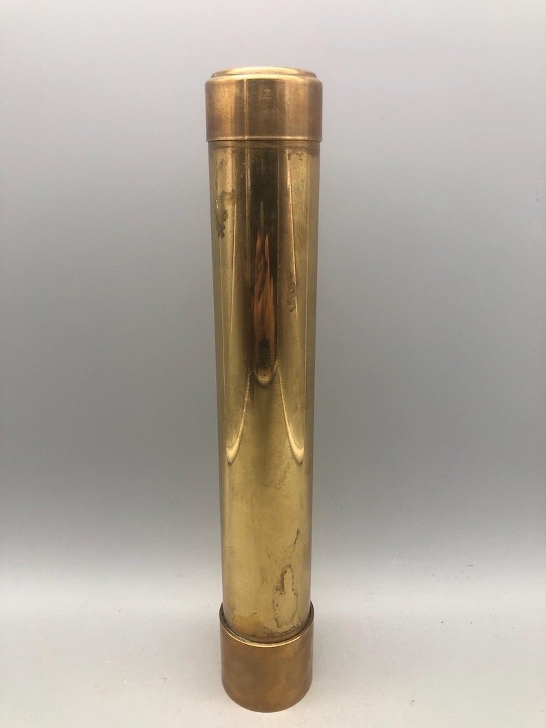 May include: A cylindrical brass container with a removable lid and base. The container has a polished, reflective surface with vertical lines. The lid and base are a darker shade of brass. The container is approximately 25 cm tall.