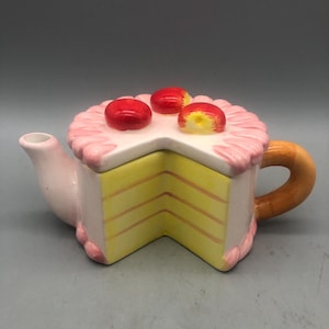 May include: A whimsical teapot shaped like a slice of cake. The teapot features a yellow cake base with pink frosting and three red cherry decorations. The handle is a light brown color, and the spout is pink.