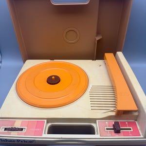 May include: Vintage Fisher-Price record player in a tan and orange color scheme. The portable player has an orange turntable, tone arm, and lid. The front panel features volume and speed controls. The Fisher-Price logo is visible.