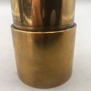 May include: Close-up of a two-piece, cylindrical brass object. The top section is slightly narrower than the bottom, with a visible seam where the two pieces connect. The brass has a polished, reflective surface.