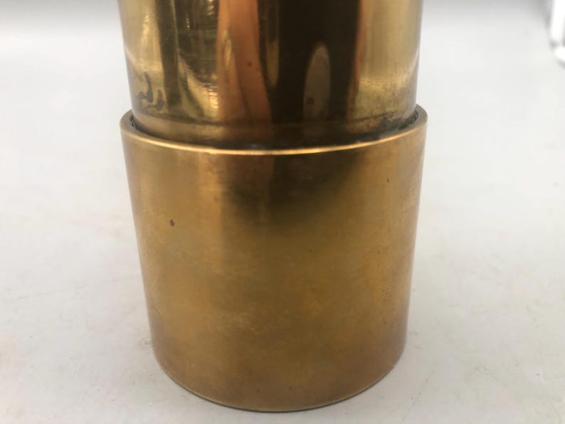 May include: Close-up of a two-piece, cylindrical brass object. The top section is slightly narrower than the bottom, with a visible seam where the two pieces connect. The brass has a polished, reflective surface.