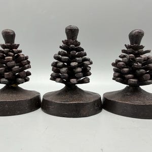 May include: Three dark brown, textured metal pinecone ornaments, each mounted on a solid, rounded base. The pinecones feature detailed, layered scales and a small, rounded tip at the top, all finished in a uniform, matte, dark brown metallic tone.