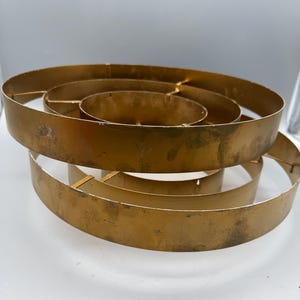 May include: A gold-colored metal structure composed of three concentric circular bands of varying diameters, connected by thin metal struts. The surface of the metal bands features a weathered, distressed finish with visible dark marks and texture.