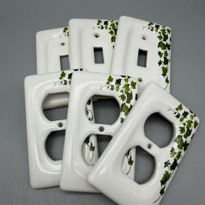 May include: A collection of six white ceramic electrical wall plates featuring a green ivy leaf pattern. Three plates are designed for single toggle switches, and three plates are designed for duplex electrical outlets, all with screw holes.