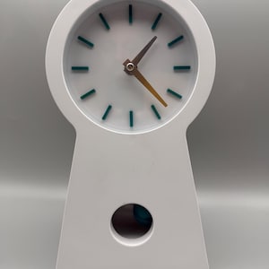 May include: A white table clock with a circular top and a tapered base. The clock face features teal hour markers and metallic hands. A circular cutout in the lower section of the clock body reveals a glimpse of a teal pendulum component inside.