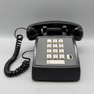 May include: A black desk telephone with a coiled cord and a push-button keypad featuring white keys with black numbers and letters. The base includes a ringer switch labeled HI, LO, and OFF, and a small rectangular label with the text 546 6291.