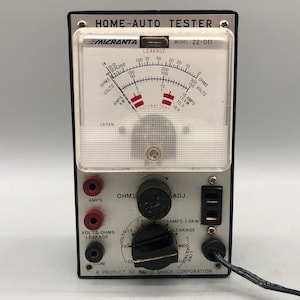 May include: A white and black Micronta Home-Auto Tester with a round analog gauge and multiple input jacks. The gauge has a red needle and a white face with black markings. The text "HOME-AUTO TESTER" is printed in black on the front of the device. The text "MICRONTA" is printed in black on the front of the device. The text "MODEL 22-011" is printed in black on the front of the device. The text "JAPAN" is printed in black on the front of the device. The text "A PRODUCT OF RADIO SHACK CORPORATION" is printed in black on the front of the device.