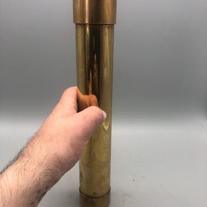 May include: A cylindrical brass object with a cap and base. The object is held by a hand. The brass has a polished finish, reflecting light. The item appears to be a container or decorative piece. The object is approximately 25 cm in length.