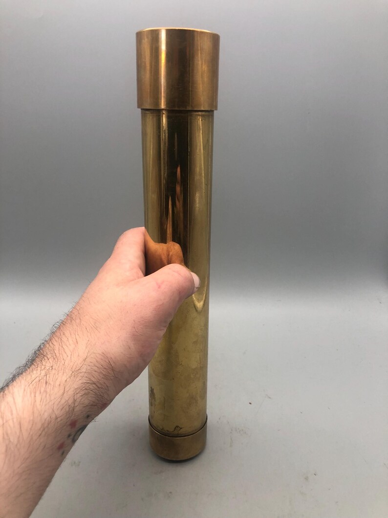 May include: A cylindrical brass object with a cap and base. The object is held by a hand. The brass has a polished finish, reflecting light. The item appears to be a container or decorative piece. The object is approximately 25 cm in length.