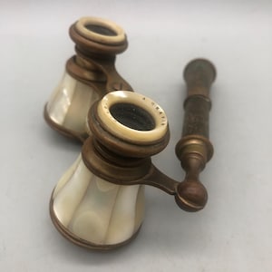 May include: Antique opera glasses with a mother-of-pearl finish and brass accents. The set includes two binoculars and a matching monocular. The lenses are surrounded by a cream-colored material, and the handles are a dark brown.