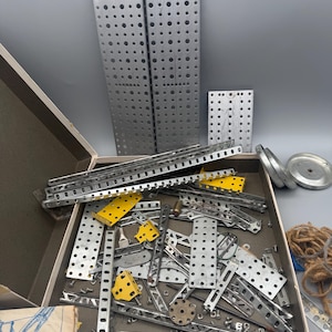 May include: A vintage metal construction set with various perforated plates, strips, and connectors. The set includes silver-colored metal pieces, some with yellow accents, and a few circular components. The pieces are stored in a cardboard box.