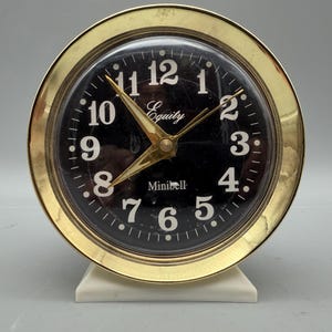 May include: A round analog alarm clock with a polished gold-tone metal frame and a black face. The clock features white Arabic numerals, gold-tone hands, and the text Equity and Minibell printed in white. It rests on a small white base.