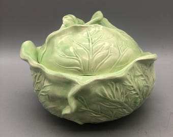 May include: A light green ceramic cabbage-shaped serving dish with a lid. The dish has detailed leaf textures and a glossy finish. The lid features a central vein pattern, and the dish is set against a gray background.