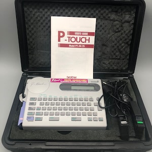 May include: A white Brother P-touch PT-20/25 label maker in a black carrying case. The label maker has a keyboard and a small LCD screen. The case has a handle and a latch.