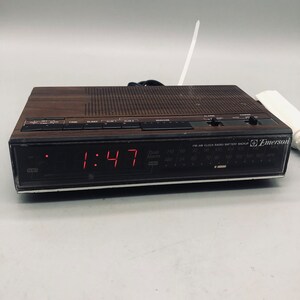 以下が含まれることがあります： 1:47 を表示するデジタルディスプレイを搭載した、茶色と黒の Emerson クロックラジオ。ラジオには内蔵アラームクロックとバッテリーバックアップ機能があります。ラジオは黒と銀のフェイスで、白いアンテナが付いています。